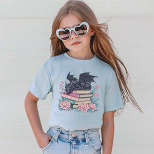 May include: A light blue t-shirt featuring a black dragon sleeping on a stack of books. The dragon is surrounded by pink and purple flowers.