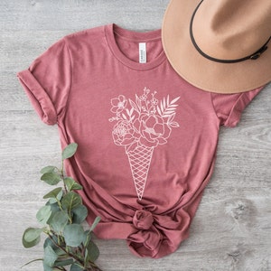 May include: A mauve t-shirt featuring a white outline of a bouquet of flowers in an ice cream cone.