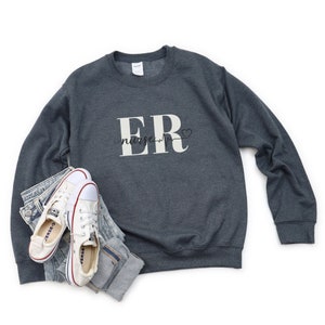 Puede incluir: Una sudadera gris oscuro con el texto "ER nurse" en blanco. La imagen tambi&eacute;n muestra zapatillas de lona blancas y vaqueros azules. El dise&ntilde;o incluye un s&iacute;mbolo de coraz&oacute;n.