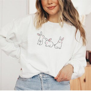 May include: White sweatshirt featuring a design of three bunnies. The bunnies are white with pink noses and are lined up.