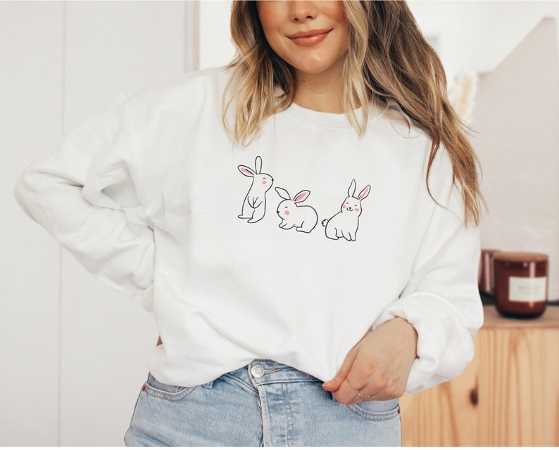 May include: White sweatshirt featuring a design of three bunnies. The bunnies are white with pink noses and are lined up.