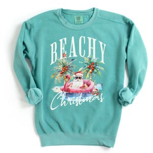 May include: Teal sweatshirt with the words "BEACHY Christmas" in white script. The graphic features Santa Claus in sunglasses, floating on a pink flamingo floatie, with palm trees and a beach scene.