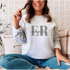 Puede incluir: Una sudadera gris claro con el texto "ER nurse" en gris. El dise&ntilde;o incluye una l&iacute;nea de latido card&iacute;aco y un s&iacute;mbolo de coraz&oacute;n. La sudadera tiene mangas largas y cuello redondo.