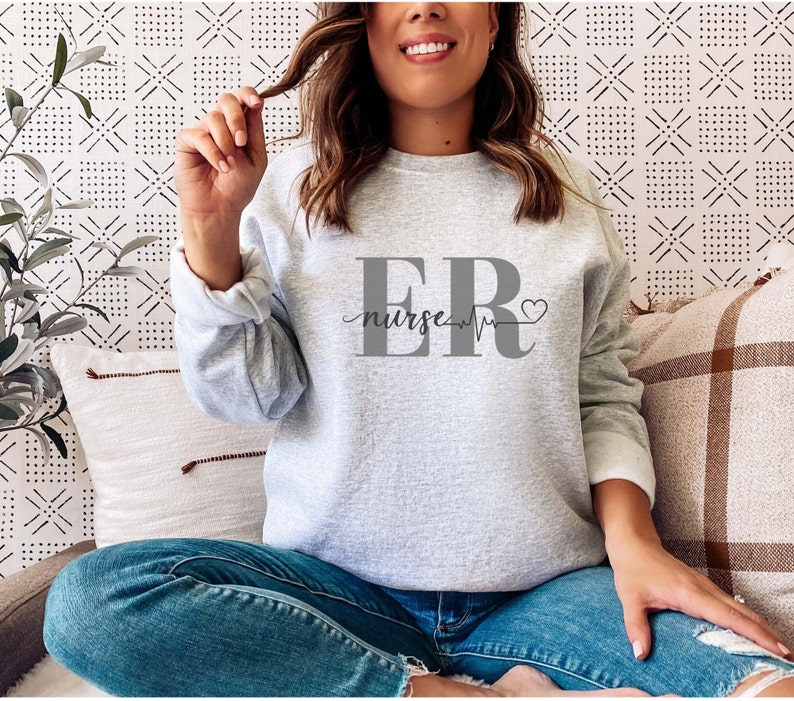 Puede incluir: Una sudadera gris claro con el texto "ER nurse" en gris. El dise&ntilde;o incluye una l&iacute;nea de latido card&iacute;aco y un s&iacute;mbolo de coraz&oacute;n. La sudadera tiene mangas largas y cuello redondo.