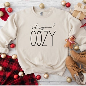 May include: A beige sweatshirt with the text "stay cozy" in black lettering. The sweatshirt is surrounded by red and gold baubles, a tartan blanket, and brown boots.