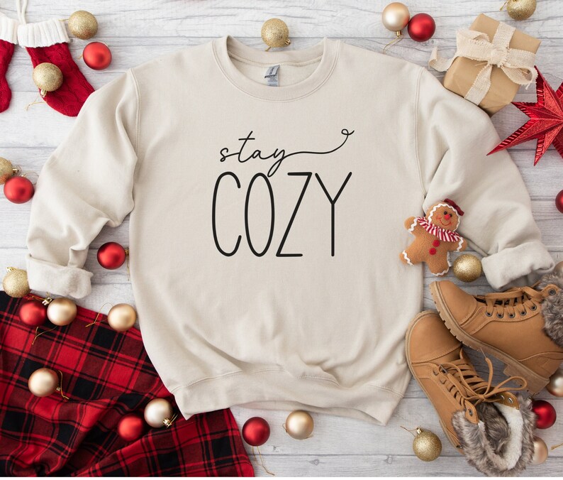 May include: A beige sweatshirt with the text "stay cozy" in black lettering. The sweatshirt is surrounded by red and gold baubles, a tartan blanket, and brown boots.