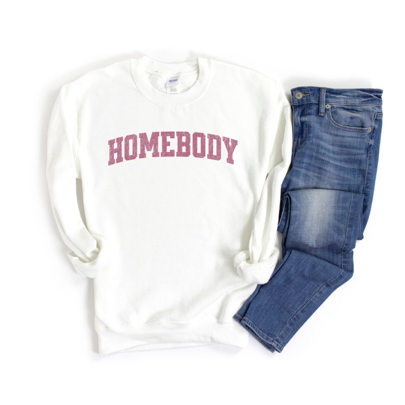 Puede incluir: Sudadera blanca con el texto "HOMEBODY" en letras rosas. La sudadera se combina con unos vaqueros azules.