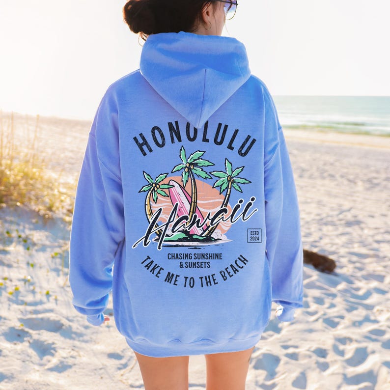 May include: Light blue hoodie with a graphic print of palm trees, a surfboard, and the text "Honolulu Hawaii" in a circle. The text "Chasing Sunshine & Sunsets Take Me To The Beach" is printed below the circle. The text "Estd 2024" is printed in a small circle to the right of the graphic.