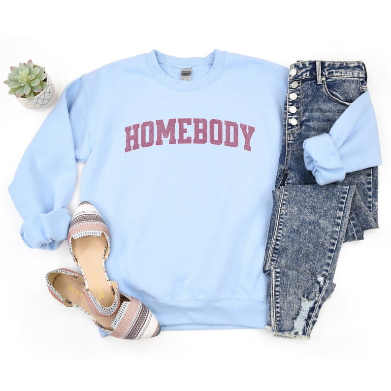 Puede incluir: Sudadera azul claro con la palabra "HOMEBODY" impresa en granate en la parte delantera. La sudadera se combina con unos vaqueros desgastados y zapatos planos estampados.
