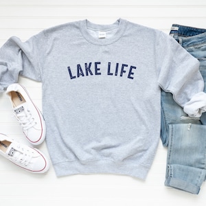 May include: A gray sweatshirt with the text "LAKE LIFE" printed in navy blue on the front. The sweatshirt is laid flat on a white wooden surface with a pair of white sneakers and blue jeans.