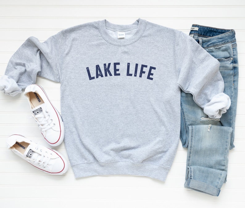 May include: A gray sweatshirt with the text "LAKE LIFE" printed in navy blue on the front. The sweatshirt is laid flat on a white wooden surface with a pair of white sneakers and blue jeans.