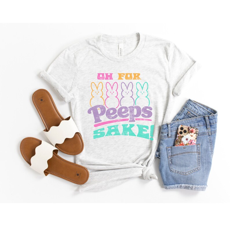 May include: A grey t-shirt with the text "Oh for Peeps Sake!" in rainbow colors. Four pink, yellow, blue, and green marshmallow Peeps are arranged in a row above the text. The shirt is laid out on a white surface with a pair of white sandals and blue denim shorts.
