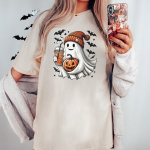 May include: A white t-shirt with a cartoon ghost wearing a brown and orange hat holding a pumpkin and a cup of coffee. The ghost is surrounded by bats and stars.