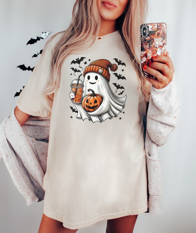 May include: A white t-shirt with a cartoon ghost wearing a brown and orange hat holding a pumpkin and a cup of coffee. The ghost is surrounded by bats and stars.