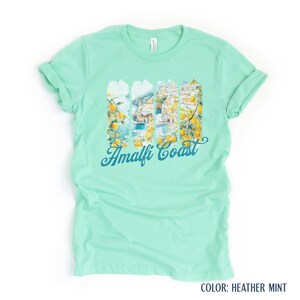 May include: A mint green t-shirt with a graphic depicting the Amalfi Coast. The design incorporates lemons, buildings, and the sea, with the words "Amalfi Coast" in a script font.