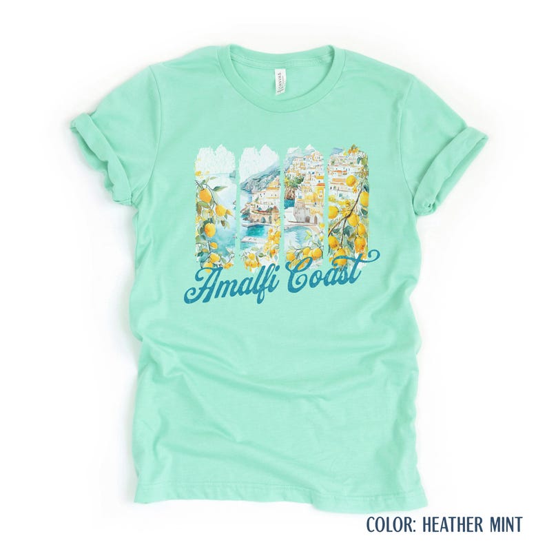 May include: A mint green t-shirt with a graphic depicting the Amalfi Coast. The design incorporates lemons, buildings, and the sea, with the words "Amalfi Coast" in a script font.