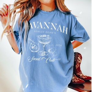 Pode incluir: Uma camiseta azul claro com um design gr&aacute;fico branco que apresenta um copo de coquetel, flores e o texto "Savannah Ashley Dean's Social Club 2024".