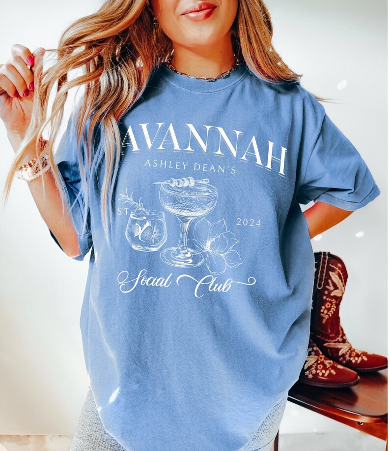 Pode incluir: Uma camiseta azul claro com um design gr&aacute;fico branco que apresenta um copo de coquetel, flores e o texto "Savannah Ashley Dean's Social Club 2024".