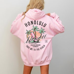 May include: Pink hoodie with a graphic print of palm trees, surfboards, and the text "Honolulu Hawaii" with the words "Chasing Sunshine & Sunsets Take Me To The Beach" below. The text "Est. 2004" is in the upper right corner of the graphic.