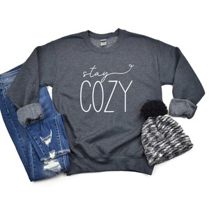 May include: A dark grey sweatshirt with the text "stay cozy" in white lettering. The sweatshirt is laid flat on a white background with a black and white knitted hat with a black pom-pom.