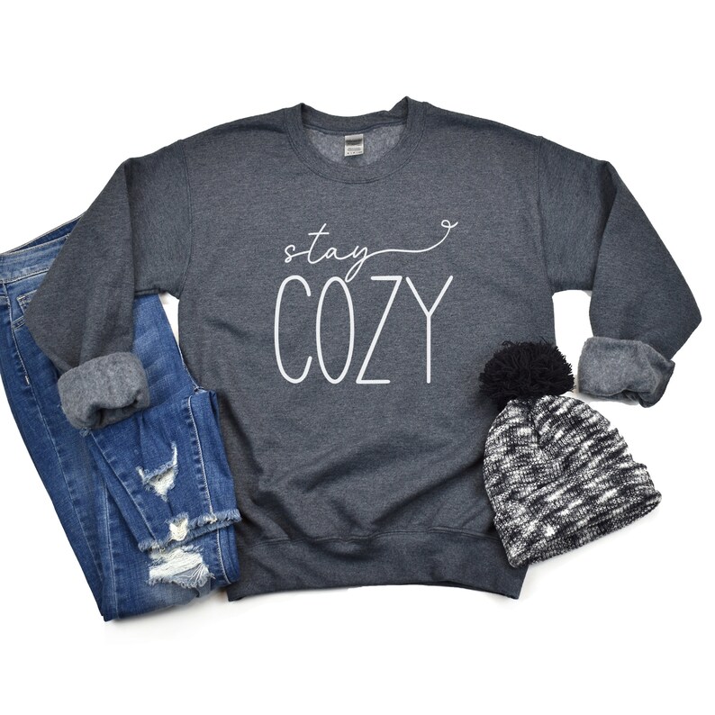 May include: A dark grey sweatshirt with the text "stay cozy" in white lettering. The sweatshirt is laid flat on a white background with a black and white knitted hat with a black pom-pom.