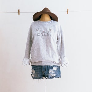 May include: A grey sweatshirt with a graphic of three bunnies on the front. The sweatshirt is on a mannequin wearing a brown hat and blue denim shorts.
