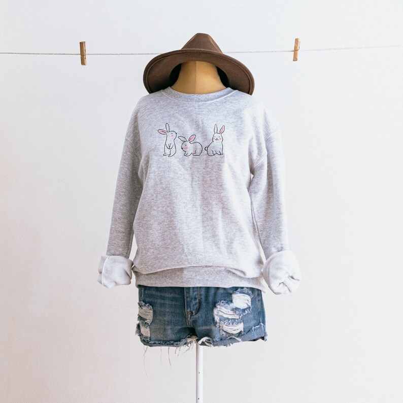 May include: A grey sweatshirt with a graphic of three bunnies on the front. The sweatshirt is on a mannequin wearing a brown hat and blue denim shorts.
