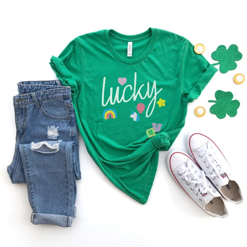 Puede incluir: Una camiseta verde con la palabra "lucky" escrita en blanco con gr&aacute;ficos coloridos de arco&iacute;ris, coraz&oacute;n, estrella y unicornio. La camiseta se combina con jeans azules y zapatillas blancas.