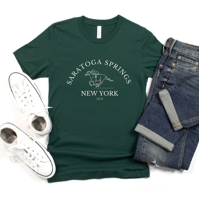 Puede incluir: Una camiseta verde oscuro con un dise&ntilde;o gr&aacute;fico blanco de un caballo y un jinete. El gr&aacute;fico incluye el texto "Saratoga Springs New York 1819".