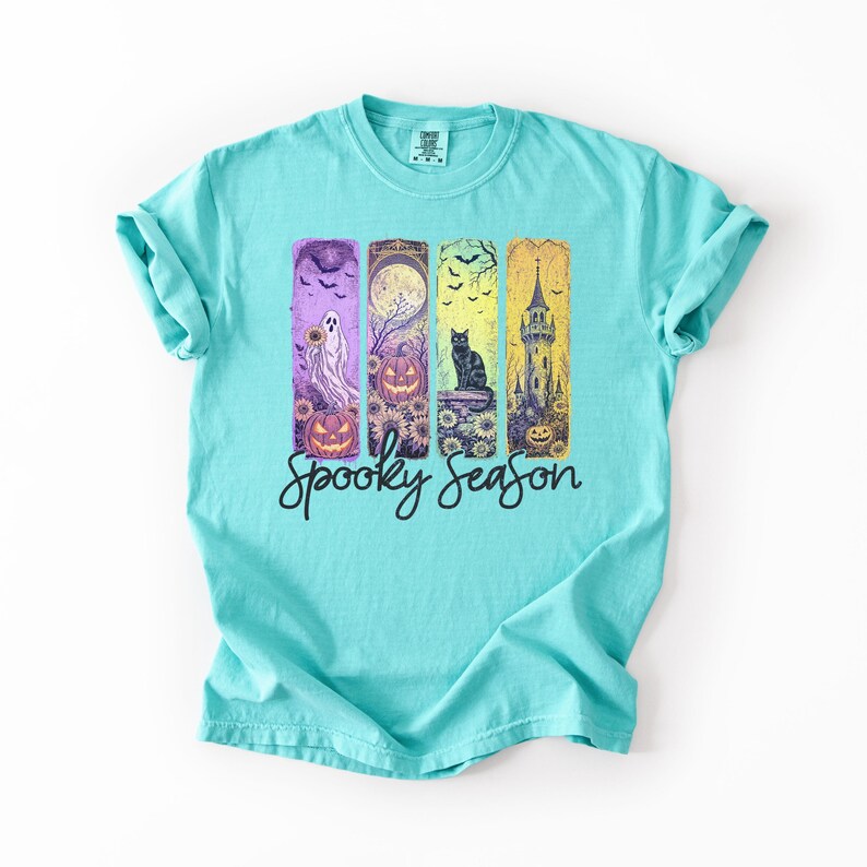May include: A turquoise t-shirt with a colorful Halloween-themed graphic. The graphic features a ghost, jack-o'-lanterns, a black cat, and a spooky castle. The words "Spooky Season" are written in a script font below the images.