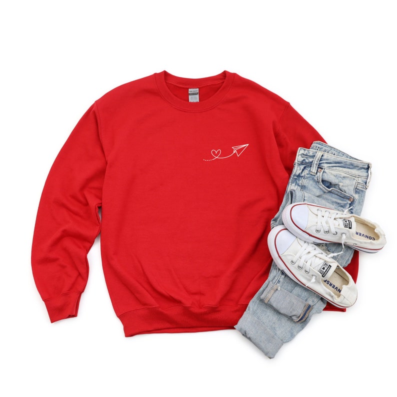 Op de afbeelding: Rood sweatshirt met een wit papieren vliegtuigje en een hartvormig ontwerp. Het sweatshirt ligt op een witte achtergrond met een paar witte sneakers en een blauwe jeans.