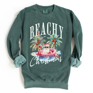 May include: A teal green sweatshirt with the words "BEACHY Christmas" in white script. The graphic features Santa Claus in sunglasses, floating on a pink flamingo in the ocean, with palm trees and red berries.