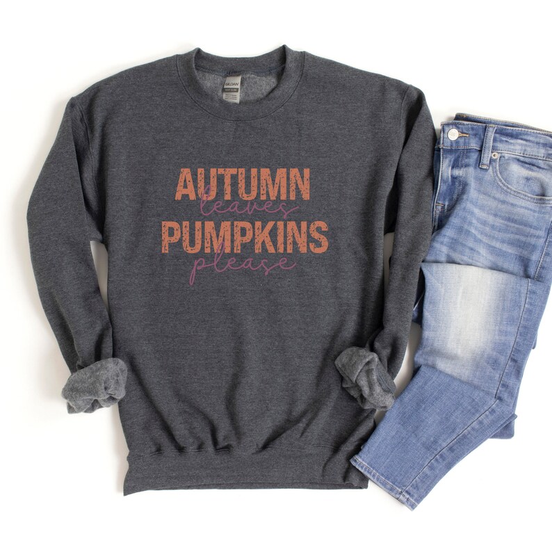 以下が含まれることがあります： オレンジとピンクで「Autumn leaves pumpkins please」と書かれた、濃いグレーのスウェットシャツ。明るいブルーデニムのジーンズと組み合わせています。デザインは秋をテーマにしています。