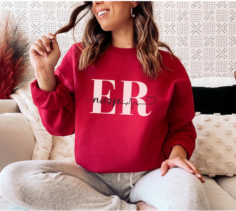 Puede incluir: Una sudadera roja con la inscripci&oacute;n "ER nurse" en blanco. El dise&ntilde;o incluye una l&iacute;nea de latido card&iacute;aco y un s&iacute;mbolo de coraz&oacute;n. La sudadera es de cuello redondo.