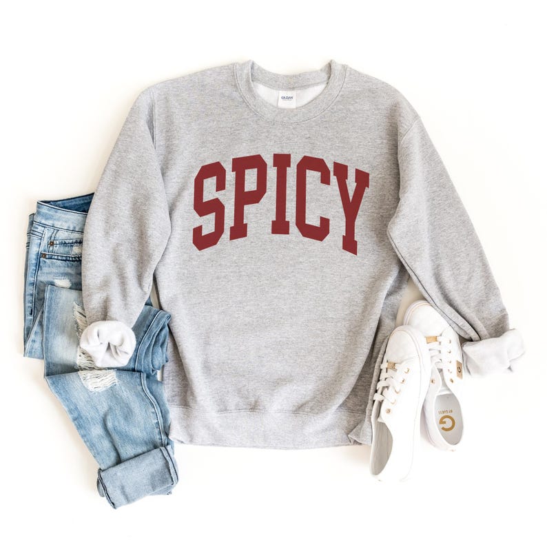 Może przedstawiać: Szara bluza z nadrukiem "SPICY" w kolorze czerwonym na przodzie. Bluza jest zestawiona z niebieskimi jeansami i białymi butami sportowymi.