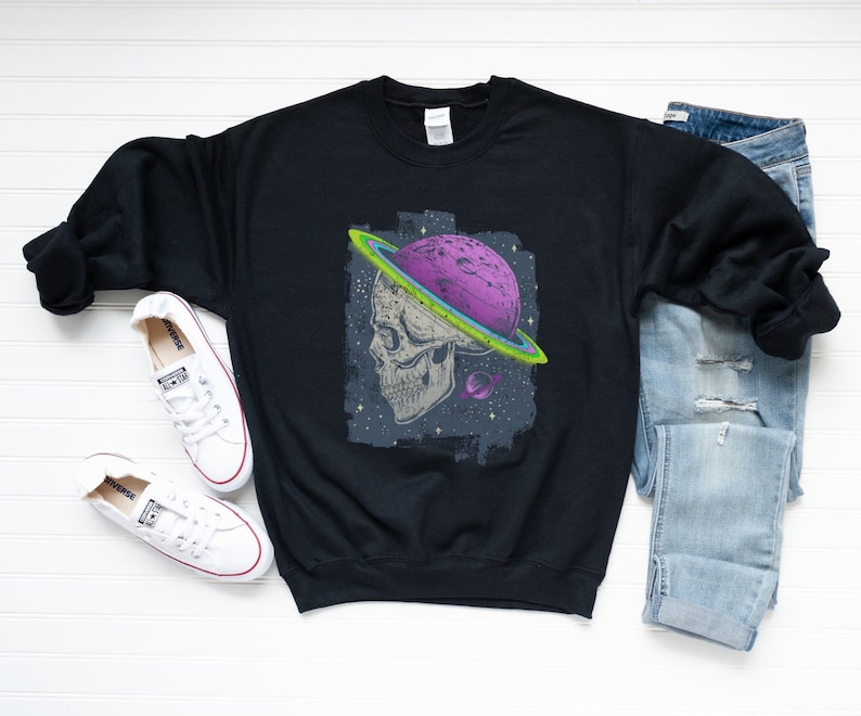 Może przedstawiać: Czarna bluza z nadrukiem graficznym czaszki z planetą i pierścieniami wok&oacute;ł niej. Planeta jest fioletowa, a pierścienie są zielone i ż&oacute;łte.