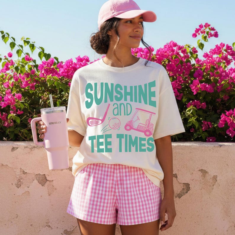以下が含まれることがあります： 「SUNSHINE and TEE TIMES」の文字とゴルフをテーマにしたグラフィックが、ターコイズとピンクでプリントされたクリーム色のTシャツ。ピンクのギンガムチェックのショートパンツとピンクのベースボールキャップを着用。大きなピンク色のタンブラーが手に持たれている。