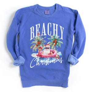 May include: A blue sweatshirt with the words "BEACHY Christmas" in white script. The graphic features Santa Claus in sunglasses floating on a pink flamingo in the ocean, with palm trees and coconuts.
