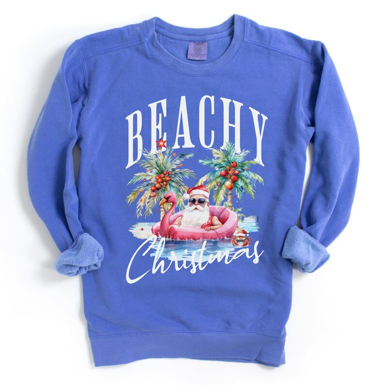 May include: A blue sweatshirt with the words "BEACHY Christmas" in white script. The graphic features Santa Claus in sunglasses floating on a pink flamingo in the ocean, with palm trees and coconuts.