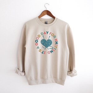 K&ouml;nnte beinhalten: Ein beiger Pullover mit einem grafischen Druck eines herzf&ouml;rmigen Wollkn&auml;uels mit Stricknadeln und dem Text "It's a good day for knitting".