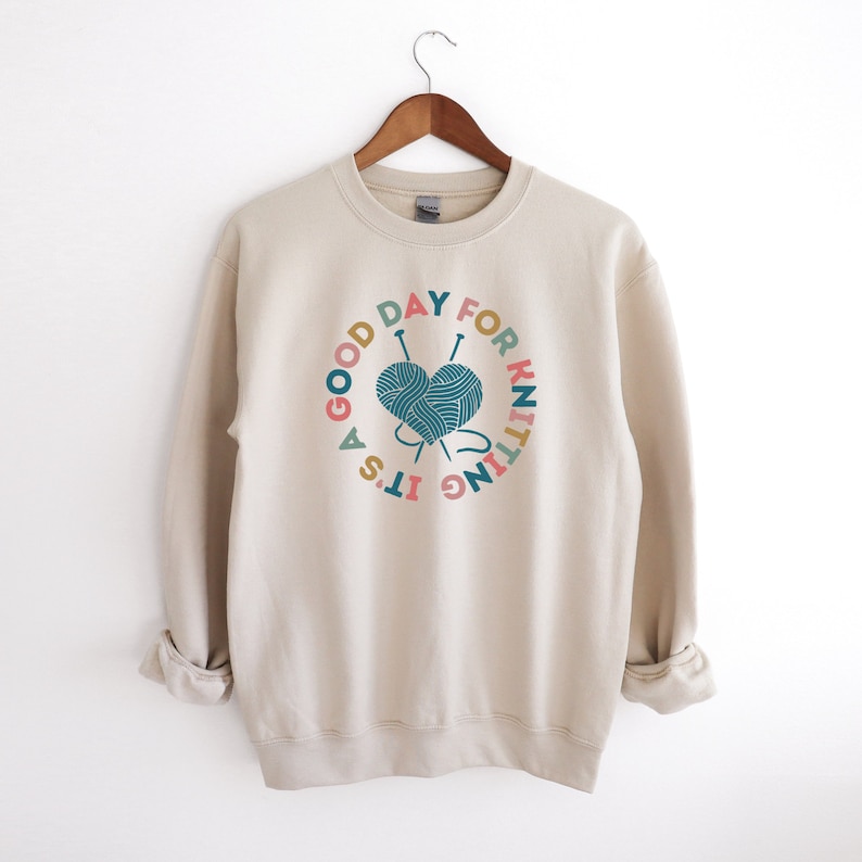 K&ouml;nnte beinhalten: Ein beiger Pullover mit einem grafischen Druck eines herzf&ouml;rmigen Wollkn&auml;uels mit Stricknadeln und dem Text "It's a good day for knitting".