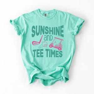 以下が含まれることがあります： 「SUNSHINE and TEE TIMES」の文字が緑色でプリントされた、明るいティール色のTシャツ。ピンク色のゴルフクラブ、ゴルフボール、ゴルフカートのデザイン。柔らかい素材のシャツです。