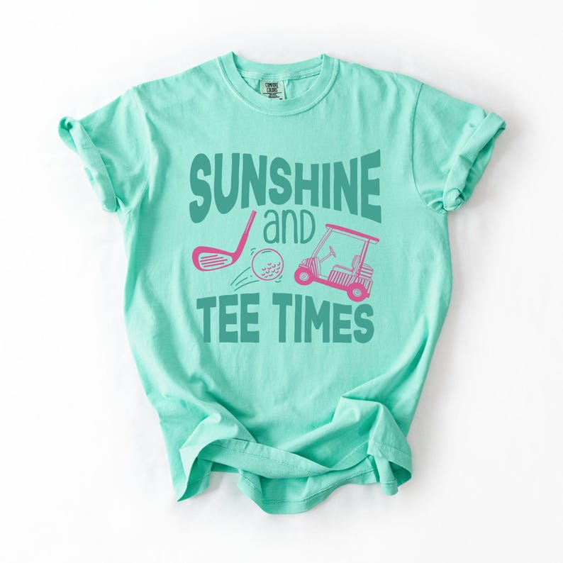 以下が含まれることがあります： 「SUNSHINE and TEE TIMES」の文字が緑色でプリントされた、明るいティール色のTシャツ。ピンク色のゴルフクラブ、ゴルフボール、ゴルフカートのデザイン。柔らかい素材のシャツです。