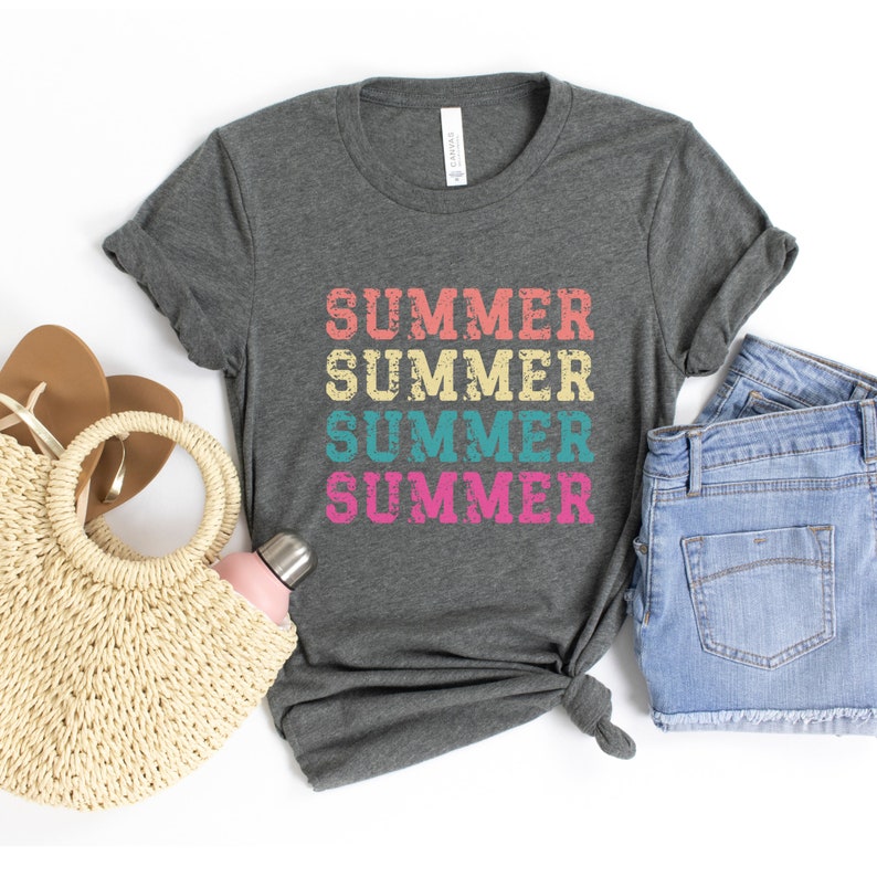 May include: Dark gray t-shirt with the word "SUMMER" printed four times in a stacked design. The word is in a distressed font with alternating colors of peach, yellow, teal, and pink. The shirt is styled with denim shorts and a straw bag.