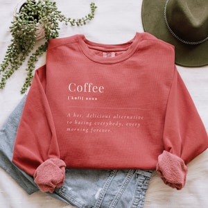 May include: A red sweatshirt with a white graphic that reads "Coffee ['kofil noun A hot, delicious alternative to hating everybody, every morning forever."
