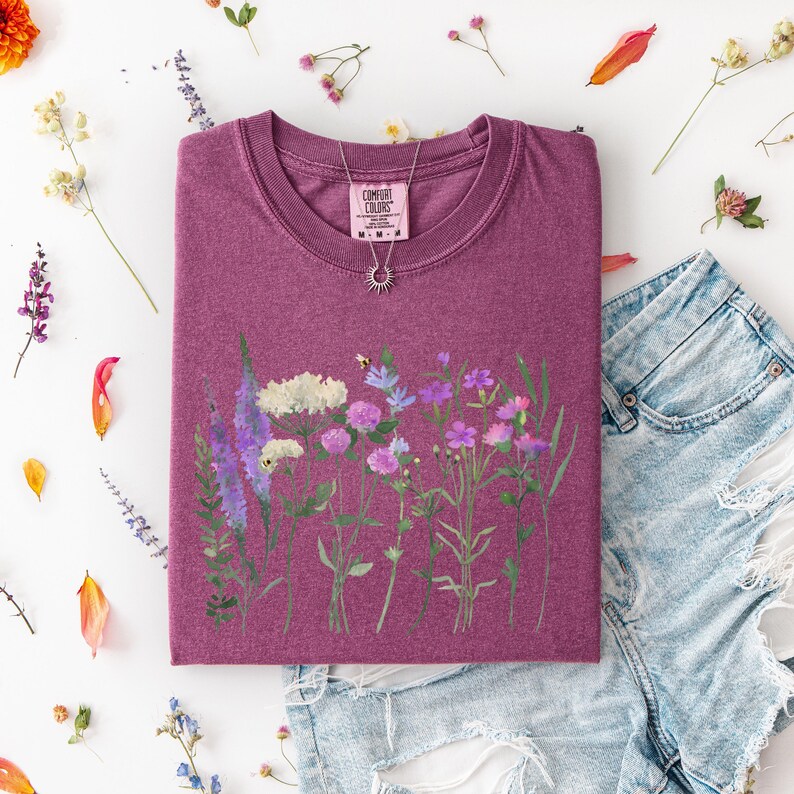 May include: A maroon Comfort Colors t-shirt featuring a watercolor floral design with lavender, white, and pink flowers. The shirt is paired with denim shorts and a silver necklace. Dried flowers and leaves are scattered around the shirt.