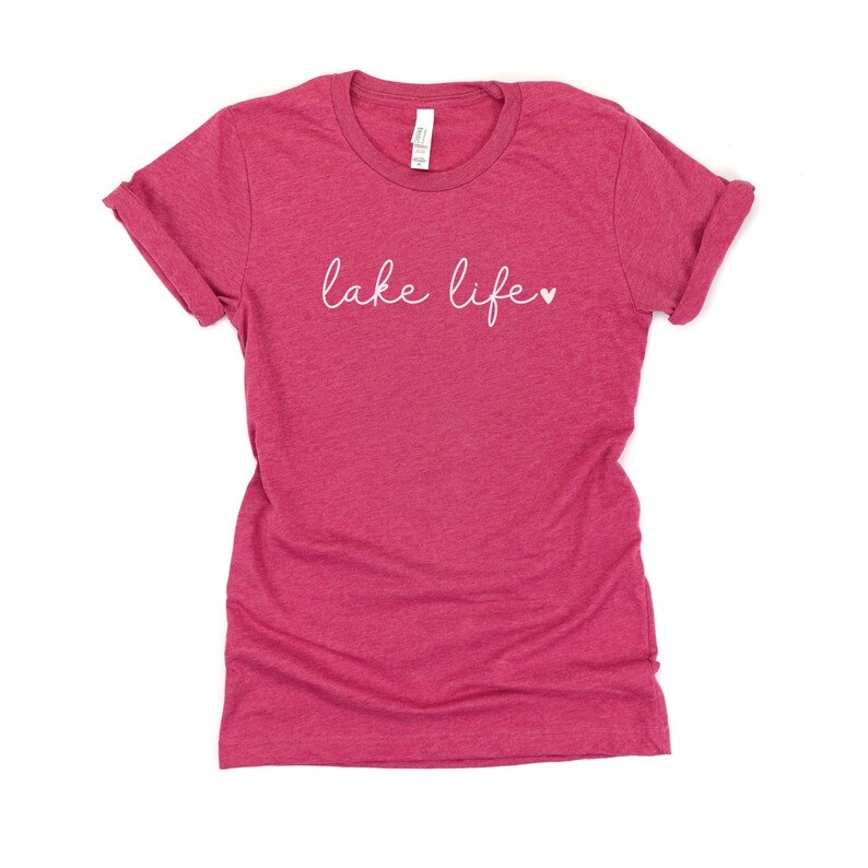 K&ouml;nnte beinhalten: Ein pinkfarbenes T-Shirt aus Heather-Material mit dem Text "lake life" in wei&szlig;er Kursivschrift und einem kleinen wei&szlig;en Herz.