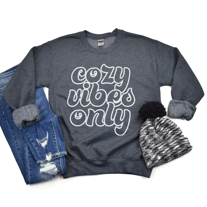 May include: A dark gray sweatshirt with the text "cozy vibes only" in white outlined letters. The sweatshirt is laid flat on a white background with a pair of blue jeans and a black and white beanie with a black pom-pom.