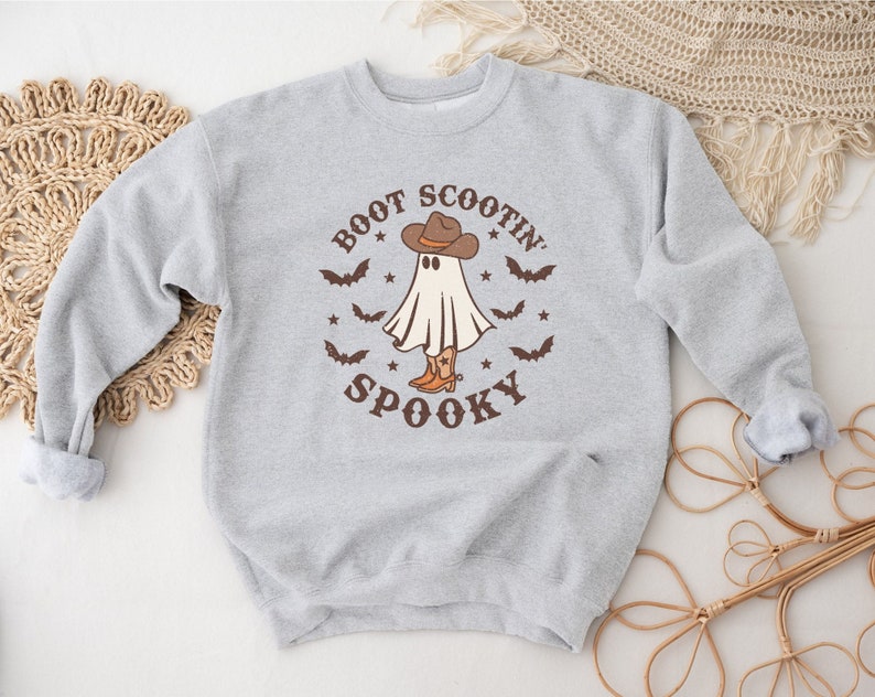 Op de afbeelding: Een grijs sweatshirt met een spook dat een cowboyhoed en laarzen draagt. De tekst "Boot Scootin' Spooky" staat op de voorkant van het sweatshirt.