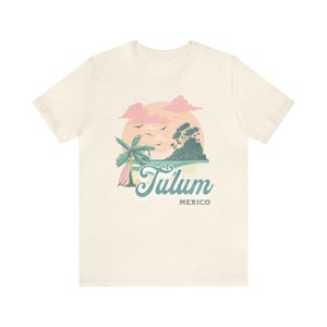 May include: A beige t-shirt with a graphic design of a palm tree, a surfboard, a small island, and a sunset. The text "Tulum Mexico" is printed below the graphic.